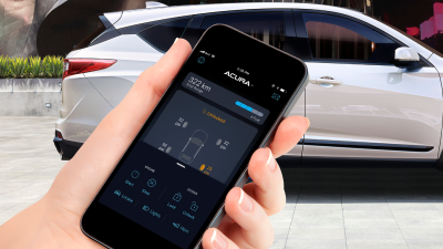 Gros plan d’une main tenant un téléphone sur l’écran duquel l’application AcuraLink est ouverte. En arrière-plan, un Acura RDX de couleur blanche est garé devant un immeuble à l’architecture moderne.