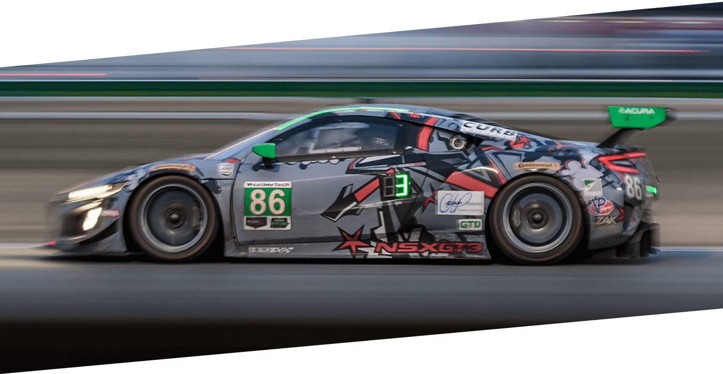 Driver side shot of a grey Acura NSX GT3 with custom sponsorship graphic wrap, driving on a racetrack with the track’s barrier blurred by the high-speed motion. // Vue du côté conducteur d’une Acura NSX GT3 grise avec un habillage graphique personnalisée roulant à pleine vitesse sur un circuit de course.